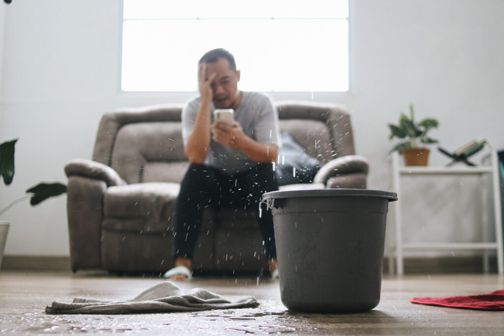 Mann sitzt verzweifelt mit Smartphone auf dem Sofa, während Wasser von der Decke tropft – ein Eimer und nasse Tücher stehen auf dem Boden im Wohnzimmer.